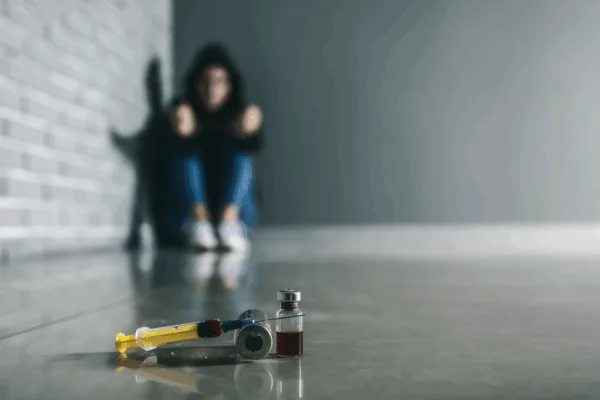 Syringes And Drug Vial On The Floor With Blurred Figure Behind, Symbolizing Substance Abuse And Mental Health Struggles.